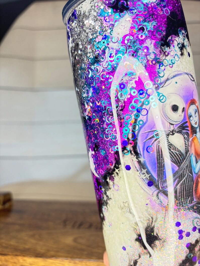 May include: A glitter-filled tumbler featuring a design from "The Nightmare Before Christmas." The tumbler has a black lid and is filled with purple, blue, and silver glitter. The design includes Jack Skellington and Sally.