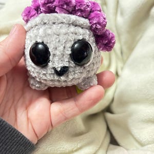 May include: A small gray crocheted plush toy with large black eyes and a purple fuzzy pom-pom on top of its head.