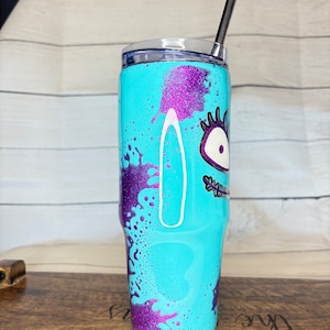 Puede incluir: Un vaso turquesa con un dise&ntilde;o de monstruo de dibujos animados. El vaso presenta detalles de purpurina morada y una tapa a juego. Una pajita negra est&aacute; insertada en la tapa. El dise&ntilde;o incluye un ojo grande y otros rasgos faciales.