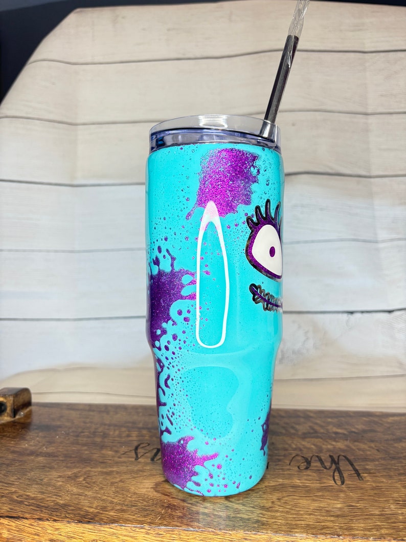 Puede incluir: Un vaso turquesa con un dise&ntilde;o de monstruo de dibujos animados. El vaso presenta detalles de purpurina morada y una tapa a juego. Una pajita negra est&aacute; insertada en la tapa. El dise&ntilde;o incluye un ojo grande y otros rasgos faciales.