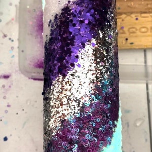 May include: A cylindrical tumbler covered in shimmering glitter. The design features a swirling pattern of white, purple, silver, and teal glitter. The tumbler is likely used for cold beverages.