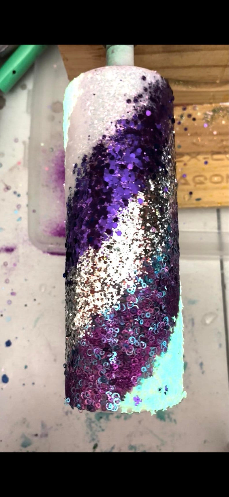 May include: A cylindrical tumbler covered in shimmering glitter. The design features a swirling pattern of white, purple, silver, and teal glitter. The tumbler is likely used for cold beverages.