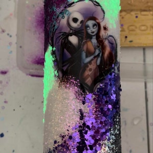 May include: A glitter-covered tumbler featuring Jack Skellington and Sally from The Nightmare Before Christmas. The tumbler has a white, purple, and black glitter design with a glowing green accent.