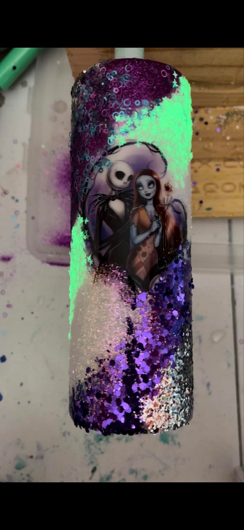 May include: A glitter-covered tumbler featuring Jack Skellington and Sally from The Nightmare Before Christmas. The tumbler has a white, purple, and black glitter design with a glowing green accent.