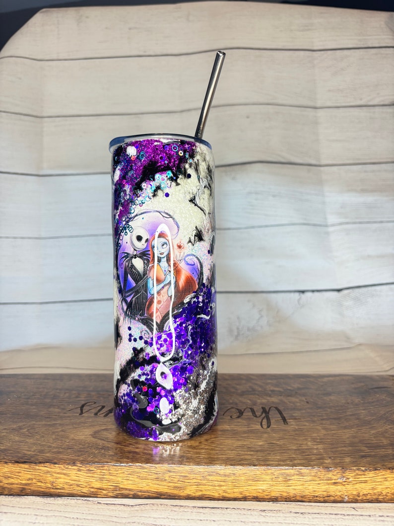 May include: A stainless steel tumbler with a black, white, and purple design featuring Jack Skellington and Sally from The Nightmare Before Christmas. The tumbler has a silver straw and is decorated with glitter.