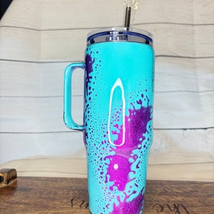 Puede incluir: Un vaso turquesa con asa, decorado con salpicaduras de purpurina morada. El vaso tiene un borde plateado y un asa turquesa a juego. Una pajita plateada est&aacute; insertada en la tapa.