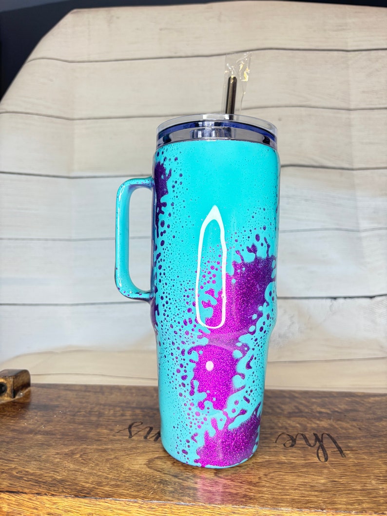 Puede incluir: Un vaso turquesa con asa, decorado con salpicaduras de purpurina morada. El vaso tiene un borde plateado y un asa turquesa a juego. Una pajita plateada est&aacute; insertada en la tapa.