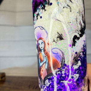 May include: A tall, cylindrical tumbler with a design featuring Sally and Jack Skellington from The Nightmare Before Christmas. The tumbler has a swirling white, black, and purple glitter design. The bottom is filled with purple glitter.