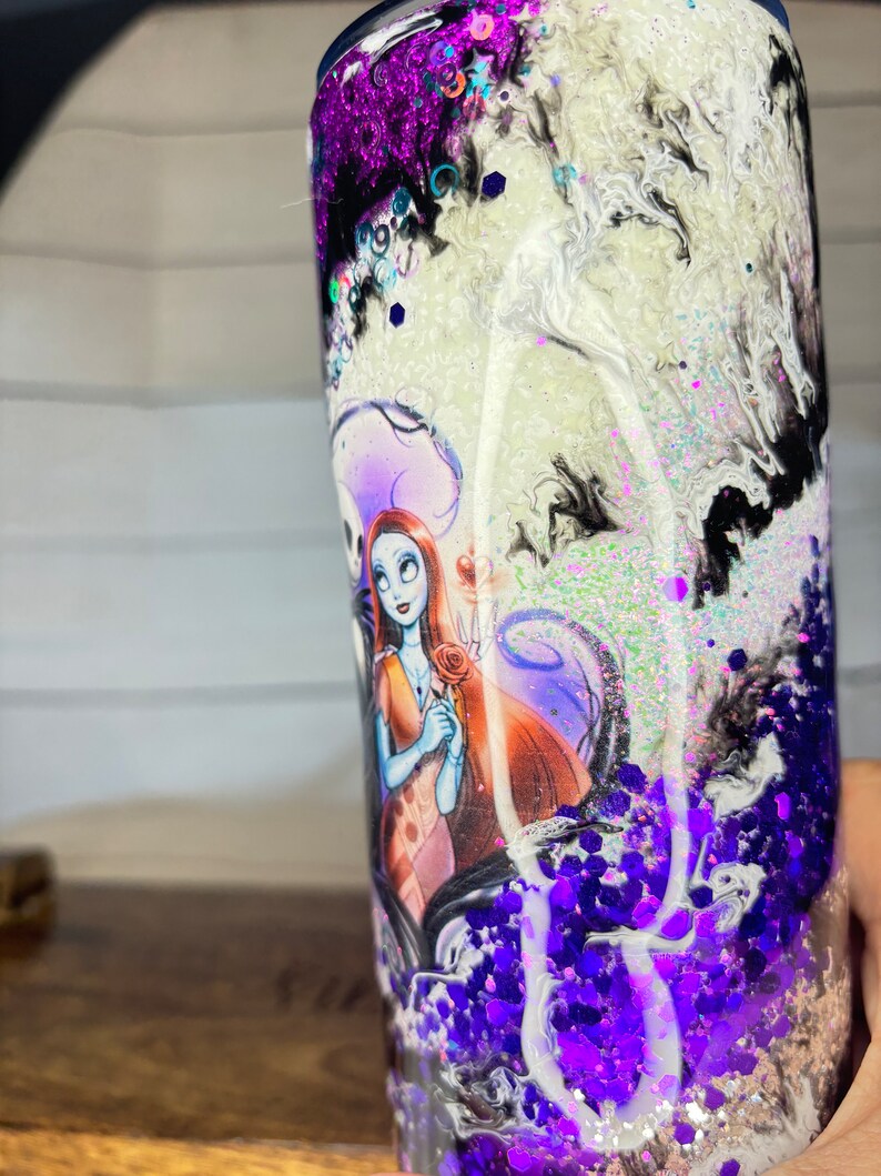 May include: A tall, cylindrical tumbler with a design featuring Sally and Jack Skellington from The Nightmare Before Christmas. The tumbler has a swirling white, black, and purple glitter design. The bottom is filled with purple glitter.