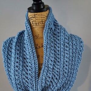 May include: A light blue crocheted scarf with a button closure. The scarf is displayed on a mannequin with a burlap-like texture.