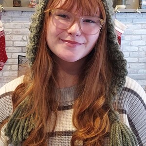 May include: A woman wearing a green crocheted hat with long tassels. The hat has a hood and covers her ears. She is wearing a striped sweater.