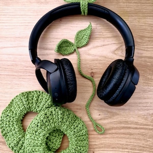 Op de afbeelding: Zwarte over-ear koptelefoon met groene gehaakte bladeren en twee groene gehaakte oorbeschermers. De bladeren zijn bevestigd aan de hoofdband. De oorbeschermers zijn cirkelvormig en getextureerd. Op een houten oppervlak.