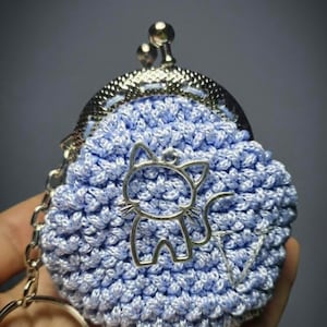 May include: A light blue crocheted coin purse with a silver-colored metal clasp and a cat-shaped charm. The purse has a silver chain and a keyring attached. The crocheted texture is visible, and the background is a gradient of gray.