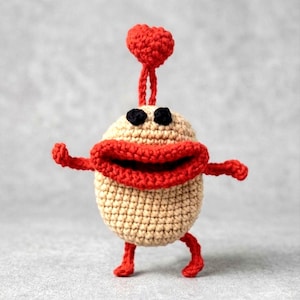 May include: A whimsical, handmade crochet creature with a beige body, red mouth, arms, and legs, and a heart-shaped headpiece. The creature has black eyes and is posed as if walking, showcasing a playful design.
