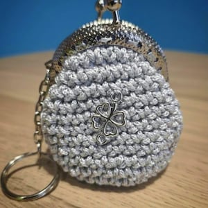 May include: A crocheted silver coin purse with a metal clasp and a keychain ring. The purse features a four-leaf clover charm and is made of a textured, hand-stitched yarn. The background is a blurred blue and brown.