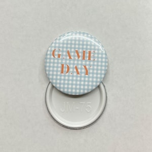 May include: A circular badge featuring a light blue and white gingham design. The words "GAME DAY" are printed in orange. The badge is partially open, showing a white metal back with the letters "JM-75".
