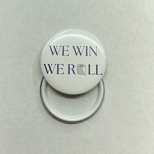 Może przedstawiać: Biały guzik z tekstem "WE WIN WE ROLL" i małym rysunkiem rolki papieru toaletowego. Tył guzika jest biały z tekstem "JMV-75".