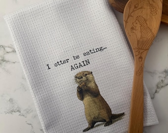 May include: White kitchen towel with a waffle weave texture, featuring the text "I otter be eating... AGAIN" and a watercolor illustration of an otter holding food. A wooden spoon with an etched design is next to the towel.
