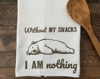 May include: White kitchen towel with a bear illustration and the text "Without my snacks I am nothing." A wooden spoon with an engraved hedgehog design is next to the towel. The towel has a textured surface.