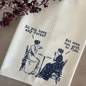 May include: White kitchen towel with a textured weave, featuring a navy blue illustration of two women and the text "Do you have any fucks?" and "Not even one, girl. Go fish."
