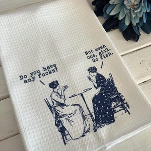 May include: White waffle-weave kitchen towel with a blue illustration of two women and text. One woman asks, "Do you have any fucks?" The other replies, "Not even one, girl. Go fish."
