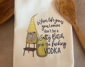 May include: White kitchen towel with a gnome illustration and the text "When life gives you lemons don't be a Salty Bitch grab the fucking VODKA." A wooden spoon with an etched design is on the right.