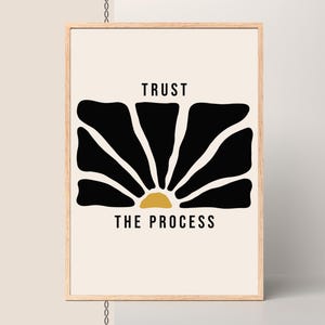 May include: A framed art print with a minimalist design. The artwork features a black floral shape with a gold sunburst center, set against a cream background. The words "TRUST THE PROCESS" are printed in black. The frame is made of light-colored wood.