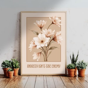 Op de afbeelding: Een ingelijste bloemenprint met een boeket witte en perzikbloemen. Het kunstwerk heeft een beige achtergrond en de tekst "ANXIETY ISN'T THE ENEMY" in bruin. Kleine potplanten staan voor het kunstwerk.