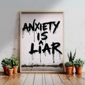 Può includere: Stampa incorniciata con il messaggio "ANXIETY IS A LIAR" in lettering nero in stile graffiti. L'opera d'arte è esposta su una parete bianca, con piccole piante in vaso davanti e un pavimento in legno.
