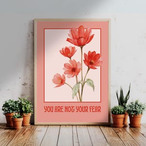 Könnte beinhalten: Ein gerahmter Kunstdruck mit einem Aquarellgemälde von roten Blumen und grünen Blättern. Das Kunstwerk ist vor einem hellrosa Hintergrund platziert und in einem hellen Holzrahmen gerahmt. Der Text "YOU ARE NOT YOUR FEAR" ist unten aufgedruckt.