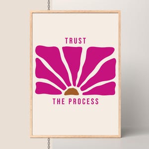 May include: A framed art print with a magenta flower design and a brown centre. The words "TRUST THE PROCESS" are printed in magenta below the flower on a cream background. The frame is made of light-coloured wood.