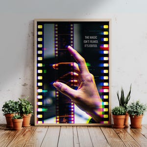 May include: Framed art print featuring a hand interacting with a film strip, with the text "THE MAGIC ISN'T FILMED. IT'S EDITED." The image has a colorful, glitch-art style. The artwork is displayed on a wooden surface.