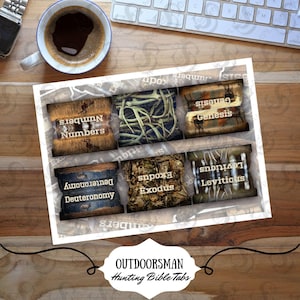 May include: A set of six Bible tabs with hunting-themed designs. Each tab features a different book of the Bible, including "Numbers," "Genesis," and "Exodus." The tabs are arranged on a white background, with a coffee cup and keyboard in the background.