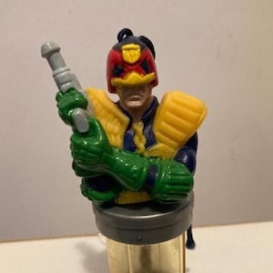 Peut inclure: Une figurine d'action en plastique de Judge Dredd, un agent des forces de l'ordre futuriste, tenant une arme à feu. La figurine porte un casque rouge, jaune et bleu et un uniforme vert et bleu. La figurine est debout sur une base en plastique transparente.