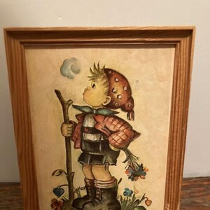 Puede incluir: Una impresión vintage enmarcada que representa a un niño con vestimenta tradicional, sosteniendo un bastón y flores. La obra de arte tiene un fondo marrón claro y está enmarcada en madera. El niño lleva un sombrero rojo y marrón y botas negras.