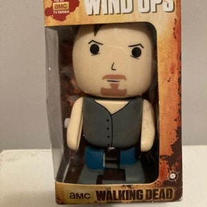 Puede incluir: Un juguete de cuerda de Rick Grimes de la serie de televisión AMC The Walking Dead. El juguete está en su embalaje original, que presenta el logotipo del programa y las palabras "Wind Ups".