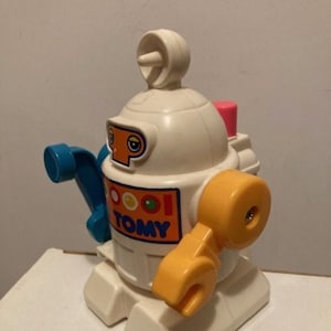 May include: A white plastic toy robot with a blue arm, yellow arm, and a pink antenna. The robot has a red and white face with black eyes and the word "TOMY" printed on its chest.