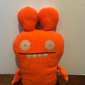May include: A bright orange plush toy with a rounded body, two tall rounded ears, and two small arms. The face features two circular beige fabric eyes with stitched details and a straight black line for a mouth with four small beige teeth.