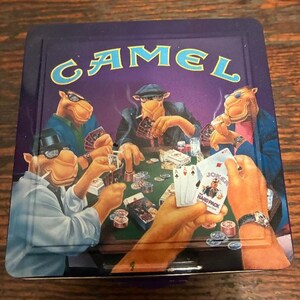 May include: A vintage Camel cigarette tin featuring a poker game scene with anthropomorphic camels. The tin has a purple background with the Camel logo in teal. The poker scene includes playing cards, chips, and cigarettes.