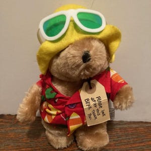 以下が含まれることがあります： 黄色の帽子と緑色のサングラスをかけた茶色のぬいぐるみクマ。赤い花柄のハワイアンシャツを着ています。クマには「このクマの世話をしてください。ありがとう。」と書かれたタグが付いています。