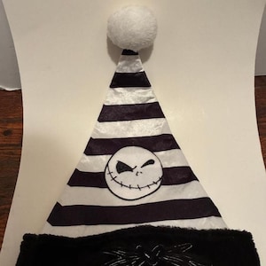 May include: A black and white striped Santa hat with a white pom-pom on top. The hat features a Jack Skellington face on the front and a black faux fur trim with a bow design. A festive holiday accessory.
