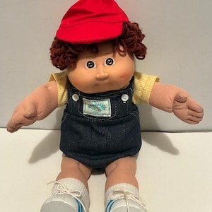 May include: A vintage Cabbage Patch Kids doll with brown curly hair, wearing a red cap, yellow shirt, and denim overalls. The doll has white trainers with blue stripes and white socks. The doll's arms are outstretched.