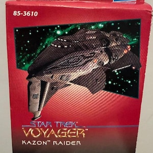 May include: A predecorated snap kit box featuring a Star Trek Voyager Kazon Raider spaceship model. The box is red with a green and black background. The model is dark gray with orange accents.