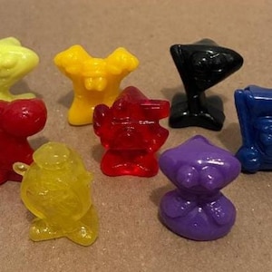 May include: A collection of eight colorful, small plastic figurines in various shapes. The figurines are in shades of yellow, red, black, blue, and purple. The figures appear to be collectible toys or game pieces.