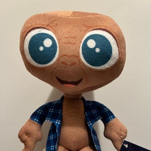 May include: A plush toy of the character E.T. The toy has large blue eyes, a tan head, and a blue plaid shirt. The toy's arms are outstretched, and it has a small smile. The toy is a collectible item.