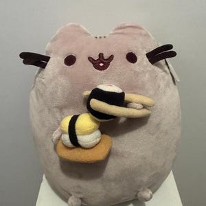 Könnte beinhalten: Ein helllila, katzenförmiges Stofftier, das zwei Sushi-Stücke hält. Das Sushi umfasst eine schwarz-weiße Rolle mit rotem Akzent und ein gelb-weißes Nigiri auf brauner Basis. Die Katze hat dunkelbraune Schnurrhaare und einen kleinen, rosa Mund.