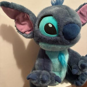 May include: A plush toy character with dark blue fur, large pointed ears with pink inner lining, and a light blue chest patch. The character features a tuft of blue hair on its head, a dark blue nose, and large eyes with light blue and black details.