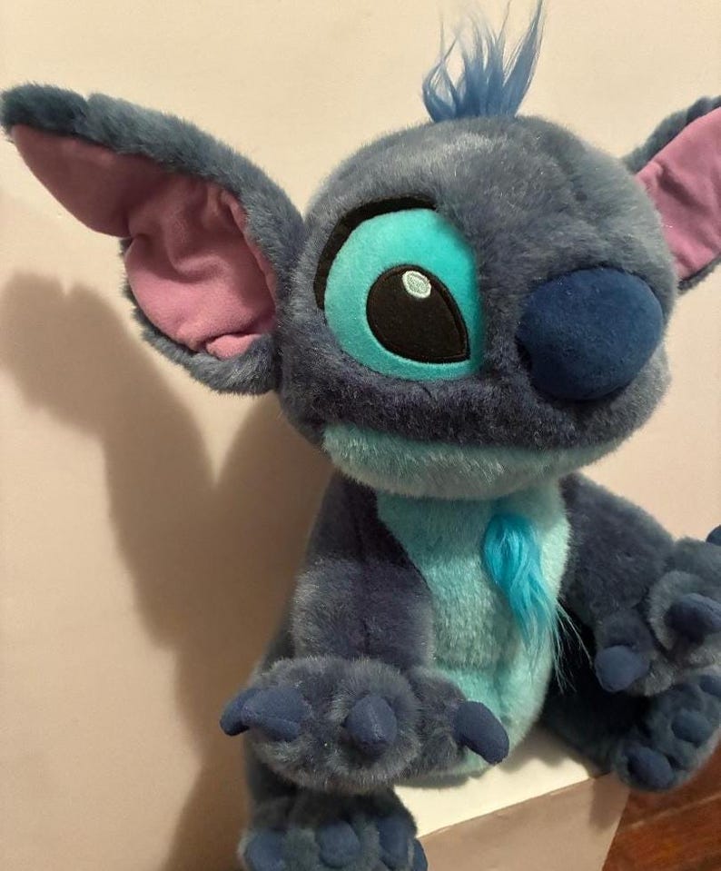 May include: A plush toy character with dark blue fur, large pointed ears with pink inner lining, and a light blue chest patch. The character features a tuft of blue hair on its head, a dark blue nose, and large eyes with light blue and black details.