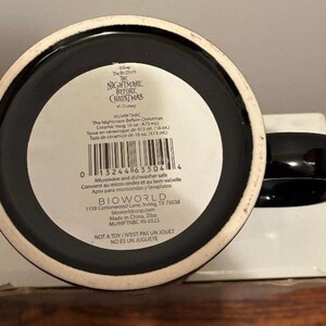 Puede incluir: Taza de cer&aacute;mica negra con asa negra y borde blanco. La base de la taza tiene una etiqueta blanca con texto que incluye "The Nightmare Before Christmas" y "Apto para microondas y lavavajillas".