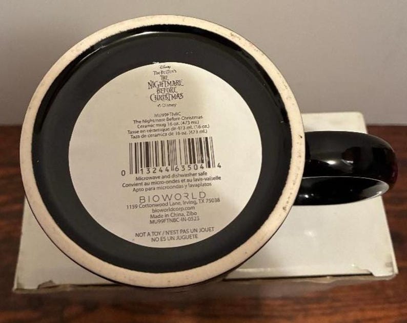 Puede incluir: Taza de cer&aacute;mica negra con asa negra y borde blanco. La base de la taza tiene una etiqueta blanca con texto que incluye "The Nightmare Before Christmas" y "Apto para microondas y lavavajillas".